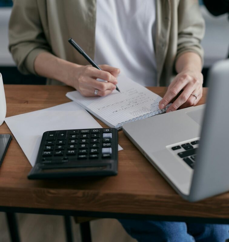 A woman writes financial calculations in a notebook, using a calculator and laptop at a wooden desk. HMRC Self Assessment Deductible Expenses